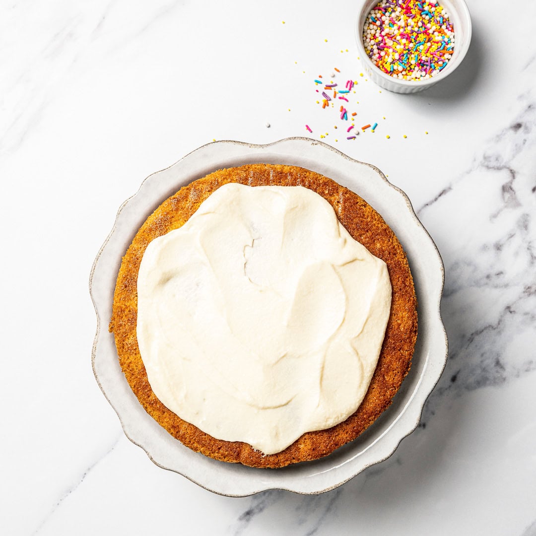 This soft and fluffy vegan vanilla cake recipe is perfect for birthdays and celebrations! It's super easy to make with pantry ingredients. Vanilla frosting spread onto layer of vanilla cake.