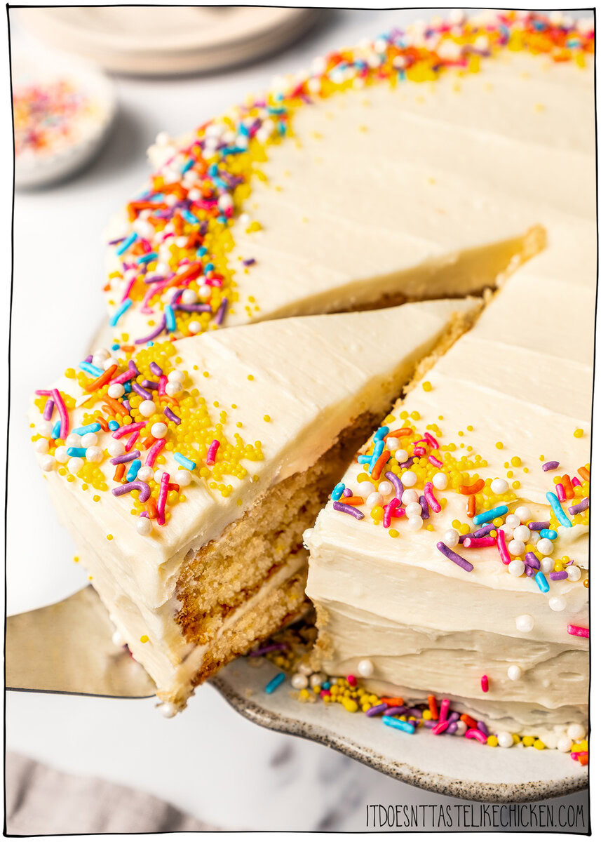 This soft and fluffy vegan vanilla cake recipe is perfect for birthdays and celebrations! It's super easy to make with pantry ingredients. Removing slice of vegan vanilla cake from platter.
