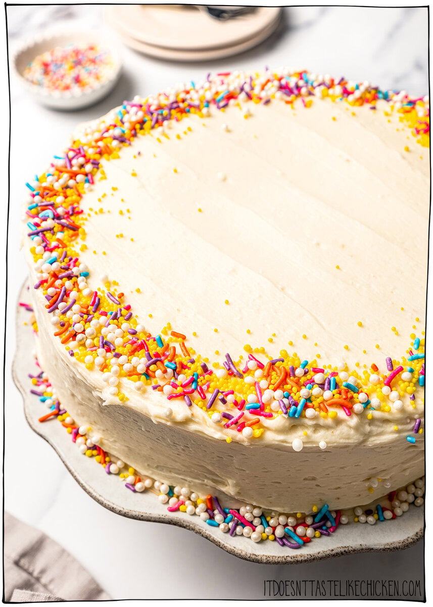 This soft and fluffy vegan vanilla cake recipe is perfect for birthdays and celebrations! It's super easy to make with pantry ingredients. Frosted vegan vanilla cake with sprinkles on platter.