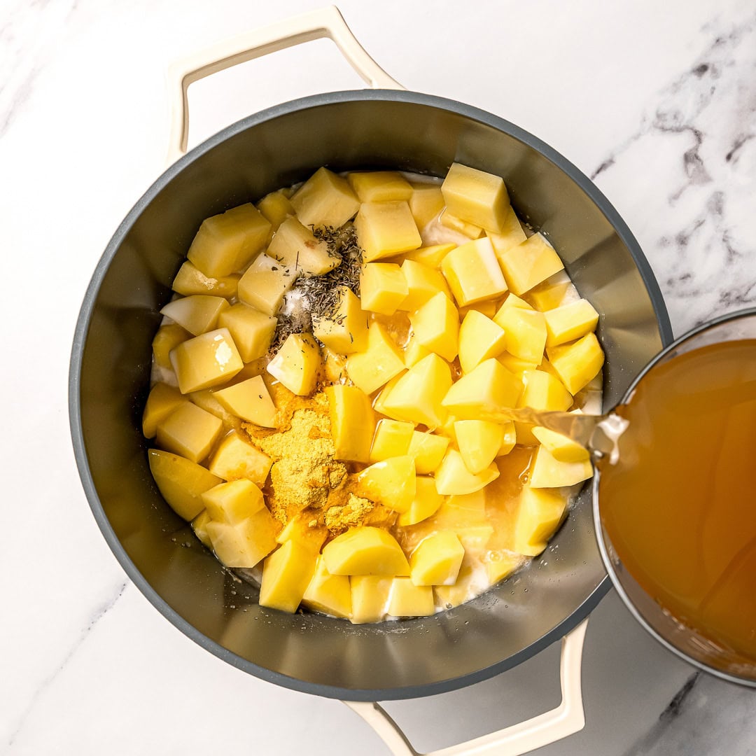 This easy creamy vegan potato soup recipe is thick, creamy, and full of cozy flavor. Think loaded baked potatoes in soup form! Pouring vegetable broth into pot of potatoes for soup.