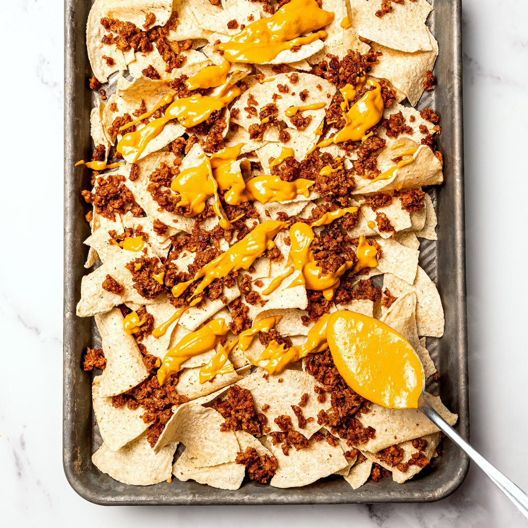 Spooning vegan nacho cheese onto tortilla chips on sheet pan.