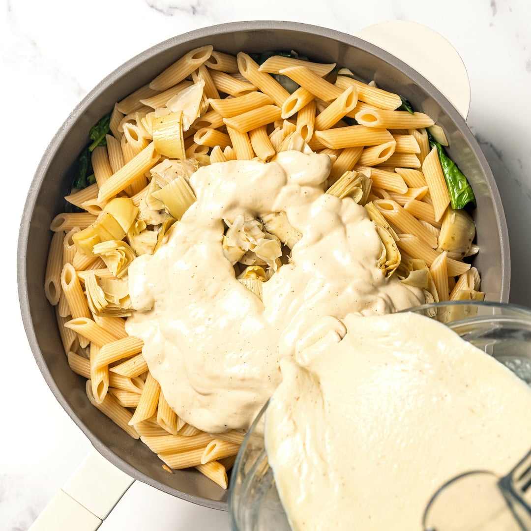 Pouring vegan cream sauce into skillet of pasta.
