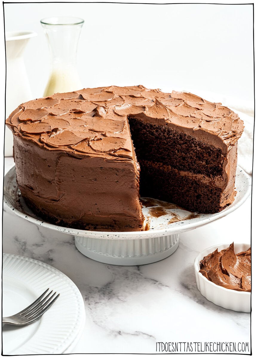 This moist, rich vegan chocolate cake is finished with fluffy chocolate buttercream, perfect for birthdays and holidays. So easy to make too! Vegan chocolate cake on cake stand with slice removed.