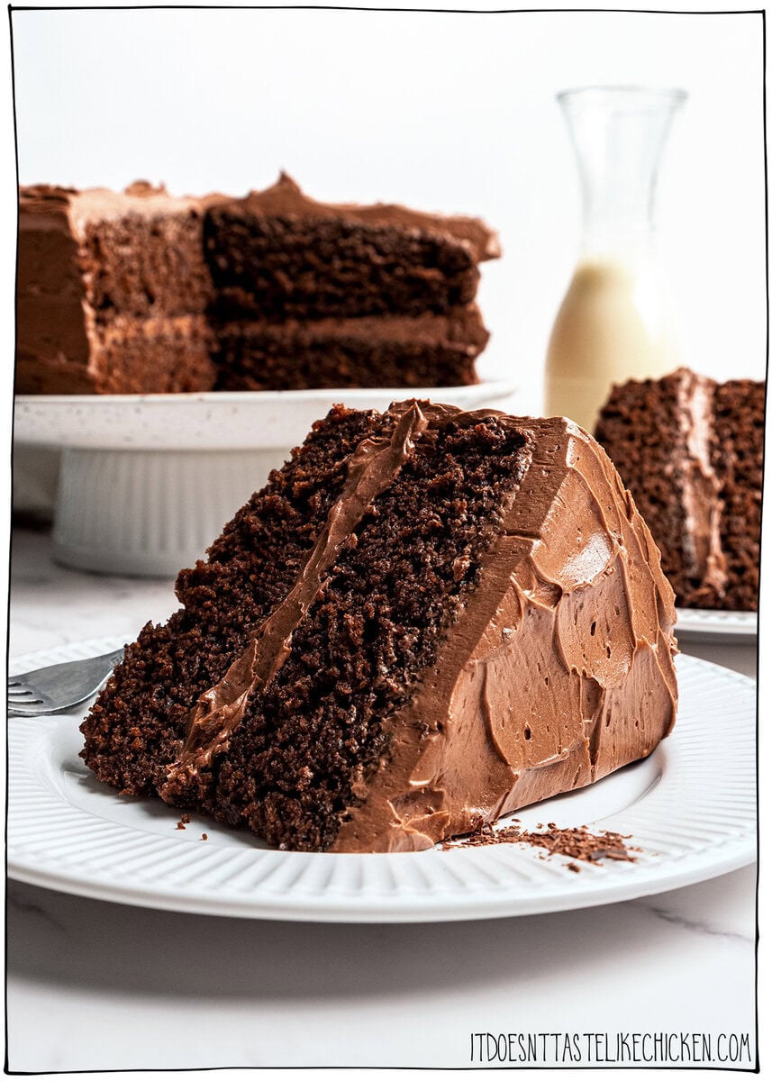 This moist, rich vegan chocolate cake is finished with fluffy chocolate buttercream, perfect for birthdays and holidays. So easy to make too! Slice of rich vegan chocolate cake on plate, with the cake on a cake stand behind.