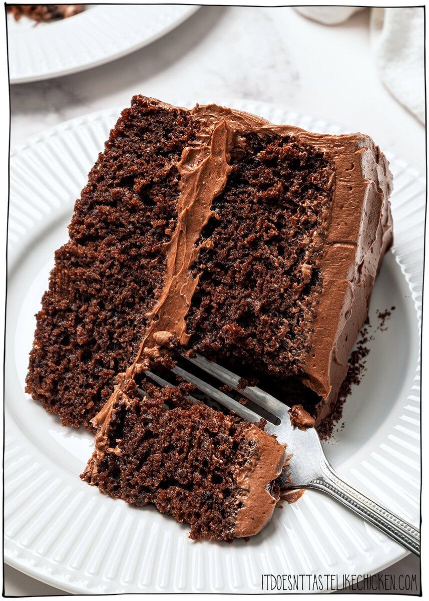 This moist, rich vegan chocolate cake is finished with fluffy chocolate buttercream, perfect for birthdays and holidays. So easy to make too! Fork cutting into vegan chocolate cake on plate.