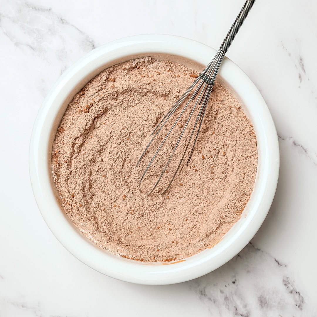 This moist, rich vegan chocolate cake is finished with fluffy chocolate buttercream, perfect for birthdays and holidays. So easy to make too! Dry ingredients for vegan chocolate cake in bowl with whisk.