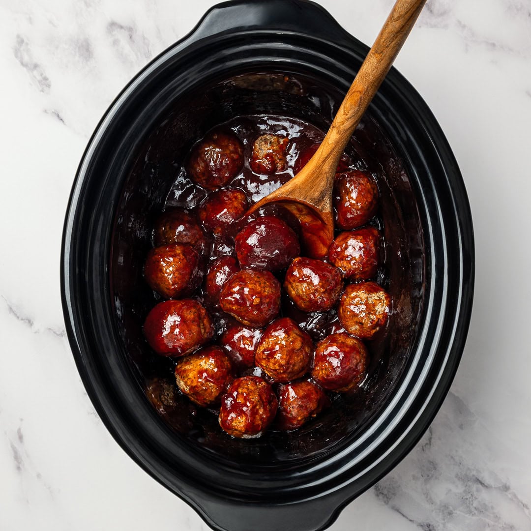 Vegan Grape Jelly Meatballs are sweet, tangy, and effortless! An easy crowd-pleasing slow cooker appetizer perfect for parties and holidays. Spoon stirring vegan meatballs into grape jelly sauce in slow cooker.