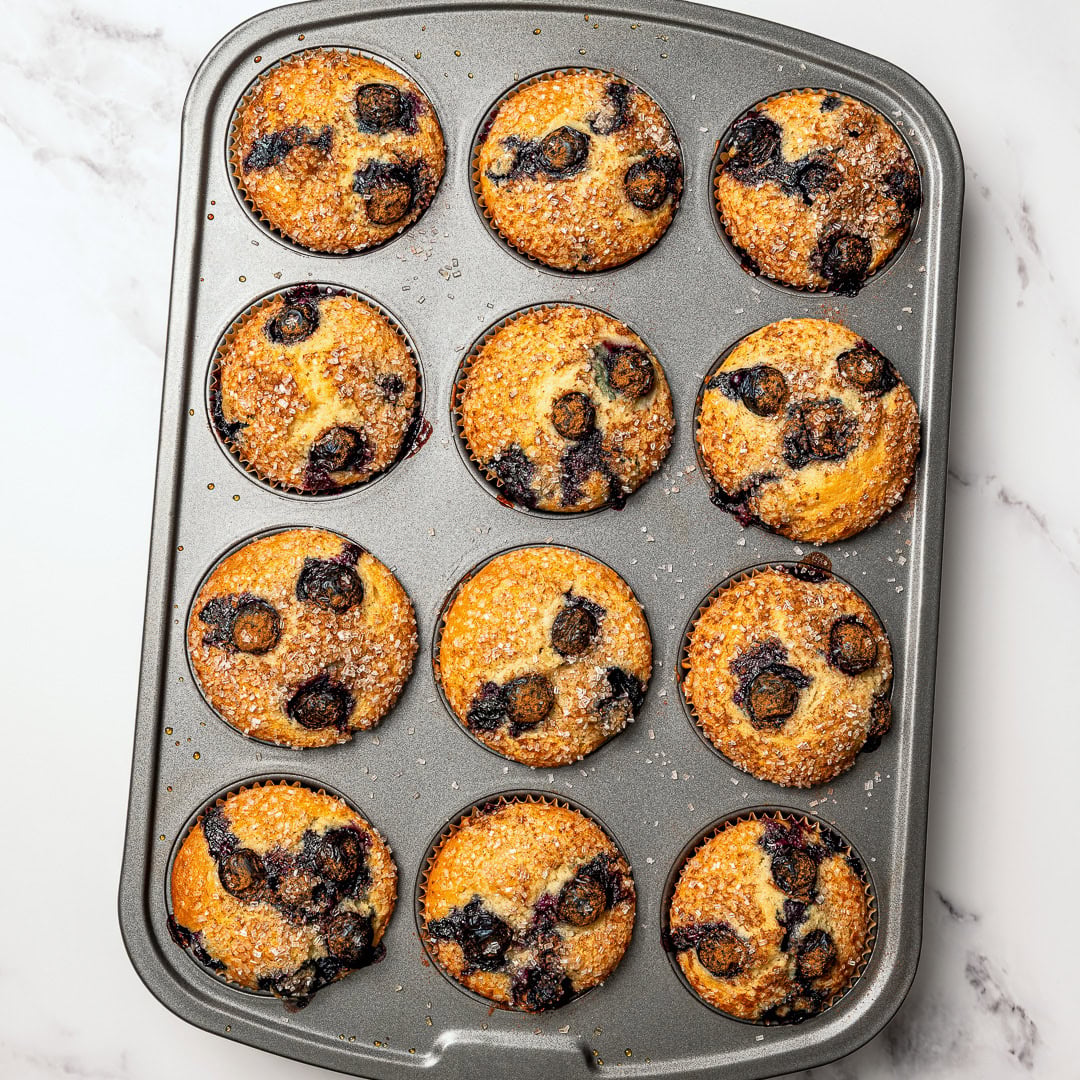 These bakery-style vegan blueberry muffins are made in one bowl, super easy! They're soft, fluffy, and finished with a crunchy sugar top. Vegan blueberry muffins in pan.