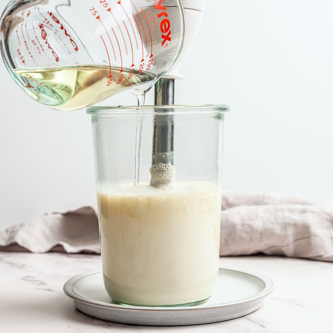 This thick, creamy vegan mayo is made in 5 minutes with soy milk! It's the BEST fool-proof recipe, egg-free, and better than store-bought. Pouring oil into jar with immersion blender making vegan mayo.