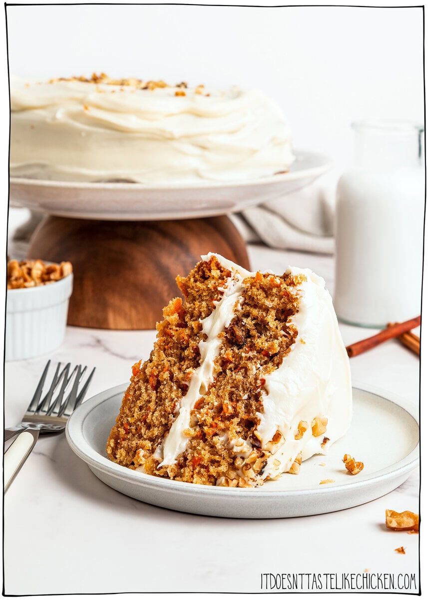 Slice of vegan carrot cake on plate with rest of cake in background.