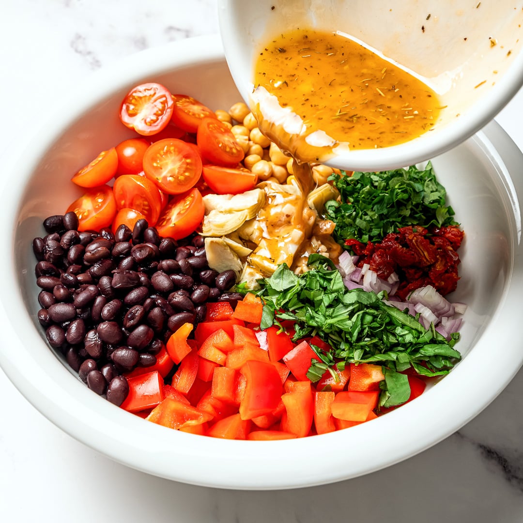 Dense bean salad packs hearty beans, crunchy veggies, and bold dressing into a satisfying make-ahead dish that stays tasty all week long! Pouring dressing into bowl of veggies for dense bean salad.