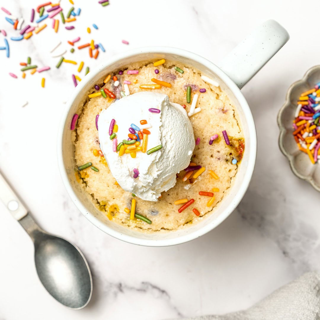 Soft, fluffy, and ready in just 5 minutes, this vegan vanilla mug cake is the perfect single-serve dessert when a sweet craving hits! Vegan vanilla mug cake with rainbow sprinkles and scoop of vegan ice cream on top.