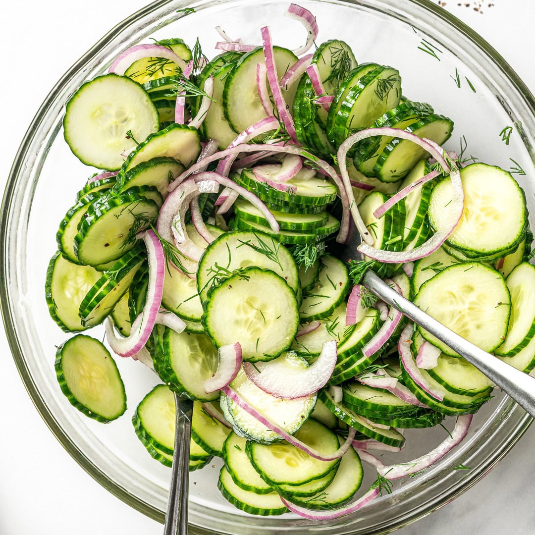 This easy cucumber dill salad is light, crisp, and refreshing, made with thinly sliced cucumbers, red onion, and a simple tangy vinaigrette. Cucumber dill salad in glass bowl with two spoons for tossing.