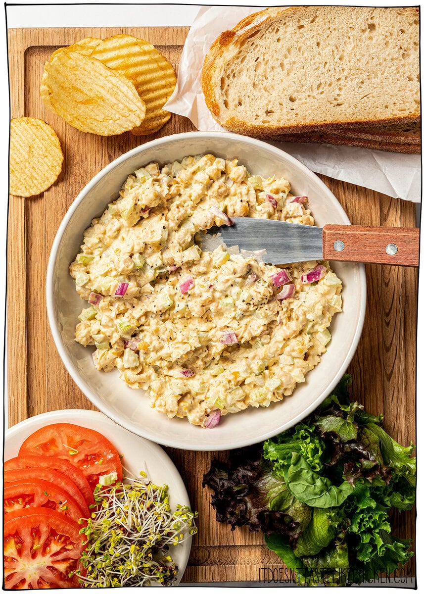 This chickpea salad sandwich is creamy, tangy and made with simple ingredients in just 10 minutes. Perfect for meal prep and quick lunches! Chickpea salad in bowl on cutting board with tomato, sprouts, lettuce, bread, and potato chips.