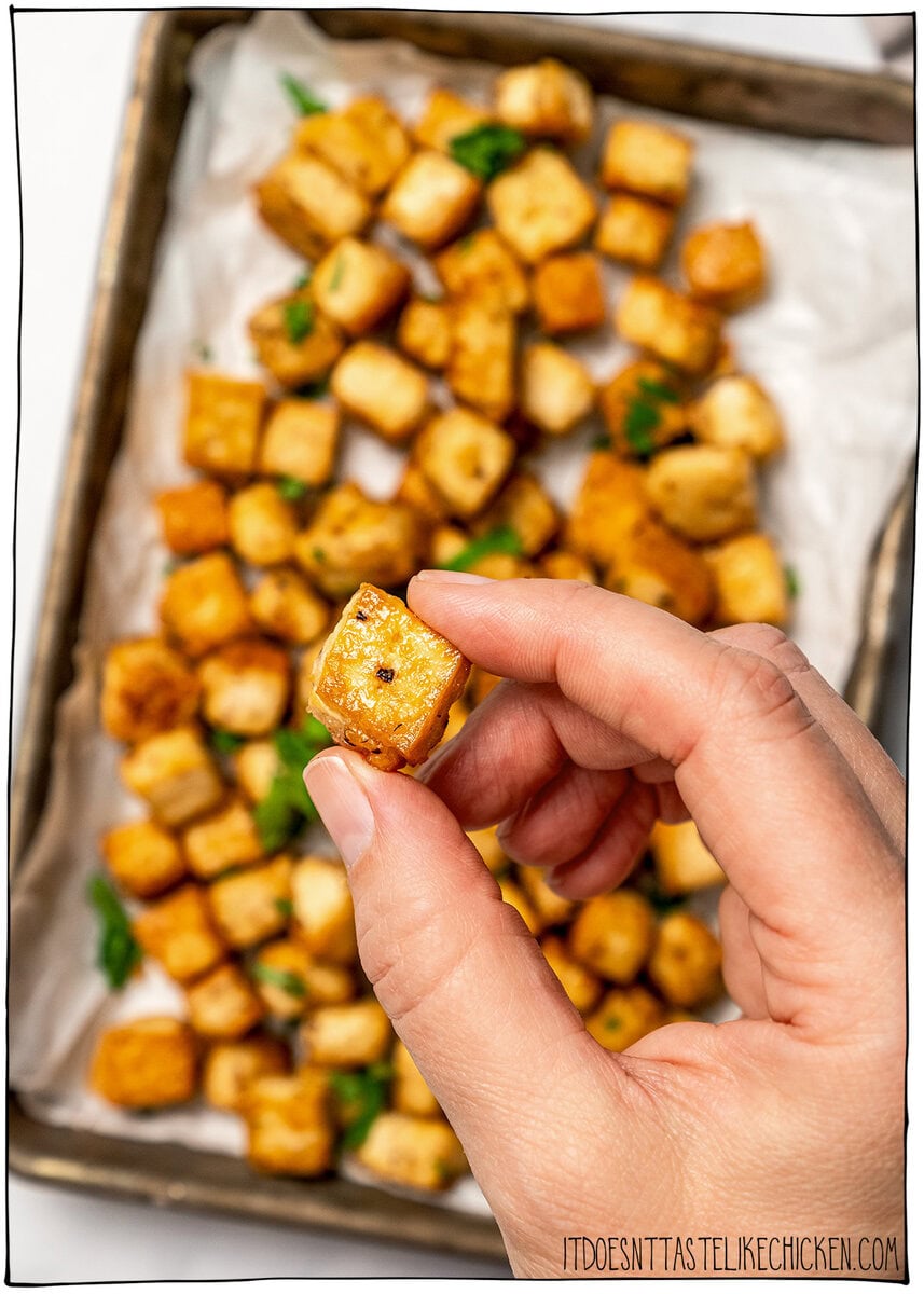 Whether you pan-fry, bake, or use an air fryer, this easy crispy tofu recipe turns out golden, flavorful, and delicious every single time! Hand holding cube of crispy tofu over sheet pan.