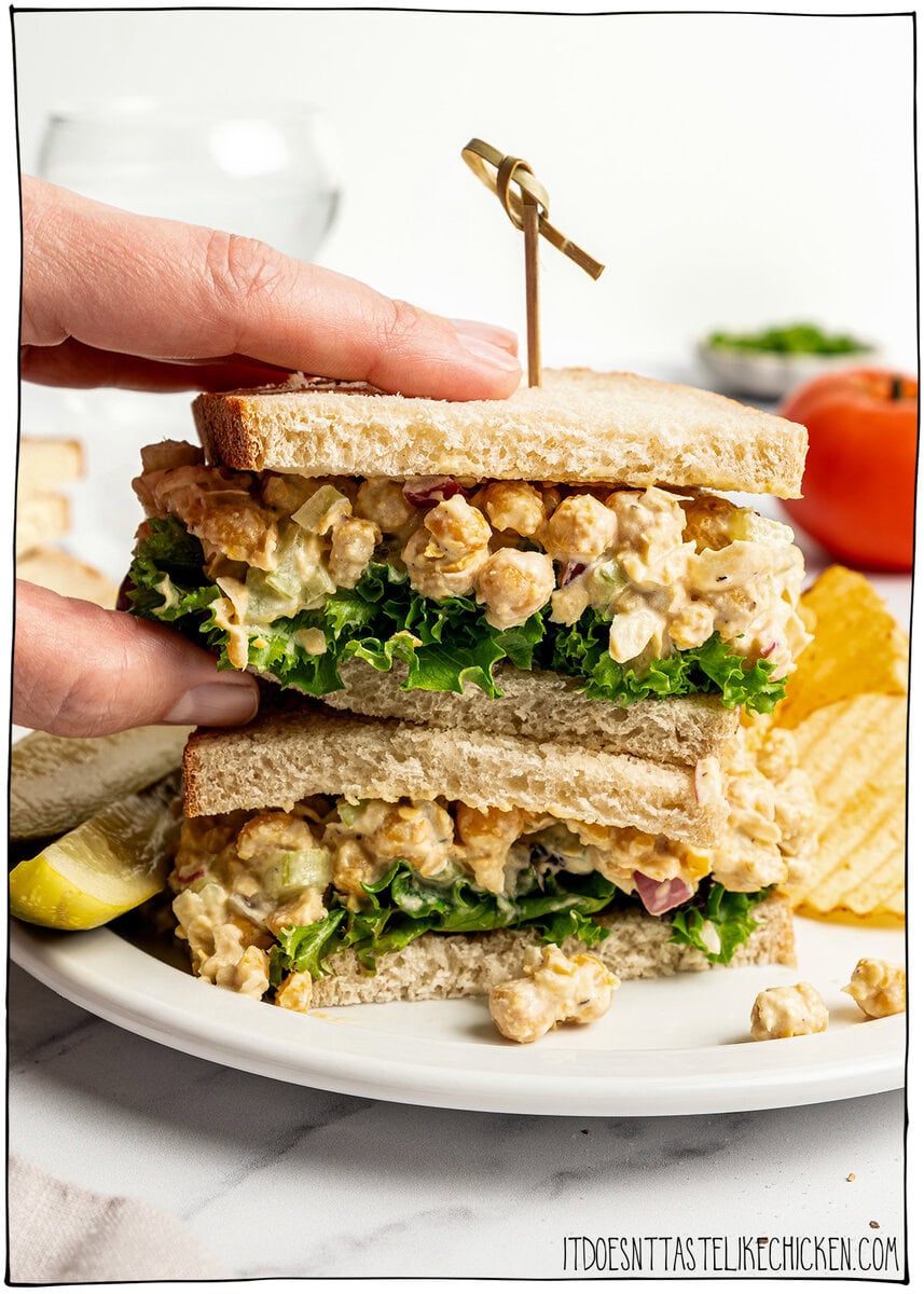 This chickpea salad sandwich is creamy, tangy and made with simple ingredients in just 10 minutes. Perfect for meal prep and quick lunches! Hand lifting chickpea salad sandwich from plate.