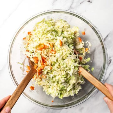 Using two wooden spoons to toss coleslaw in glass bowl.