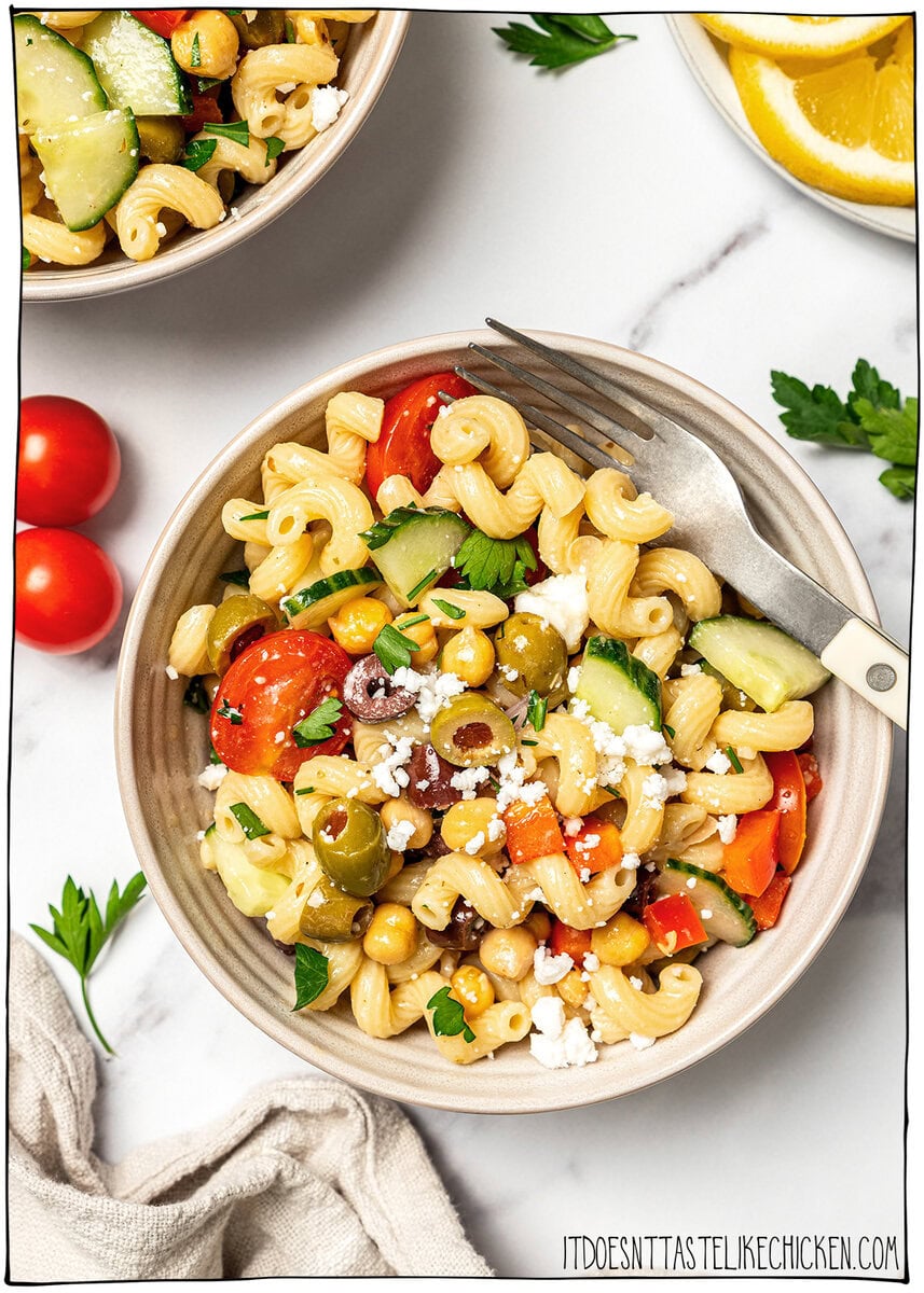Bowl of pasta salad with text overlay that reads Mediterranean chickpea pasta salad. Bowl of Mediterranean chickpea pasta salad with fresh vegetables and vegan feta.