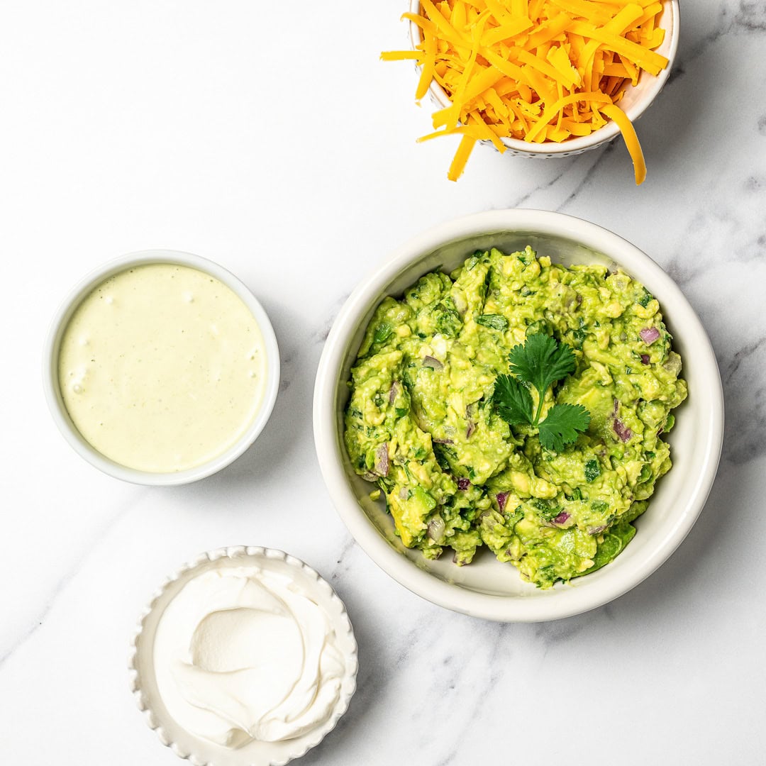 A guide to building your own burrito bowl with rice, protein, veggies, salsa, and creamy toppings for an easy and satisfying vegan meal. Four bowls with vegan cheddar cheese, guacamole, vegan sour cream, and avocado crema.