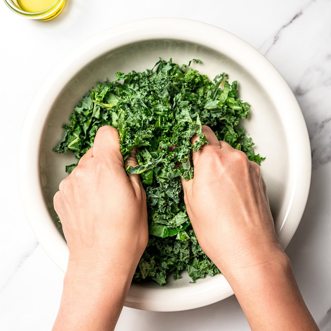 With shredded kale, zippy lemon dressing, dried cranberries, almonds and crispy chickpeas, this easy kale salad recipe is the best ever! Hands massaging kale in large bowl.