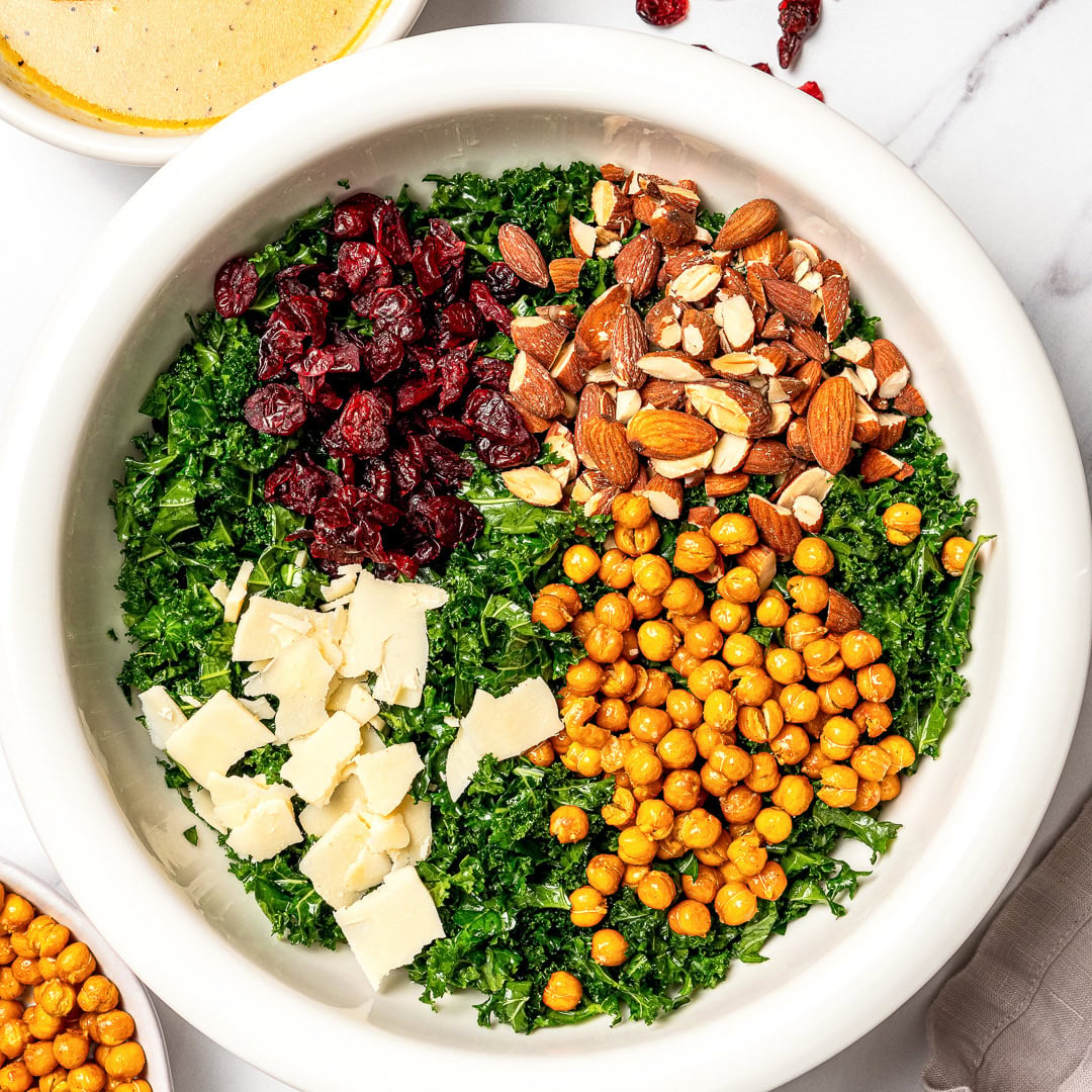With shredded kale, zippy lemon dressing, dried cranberries, almonds and crispy chickpeas, this easy kale salad recipe is the best ever! Kale, vegan Parmesan, crispy chickpeas, almonds, and dried cranberries in bowl.
