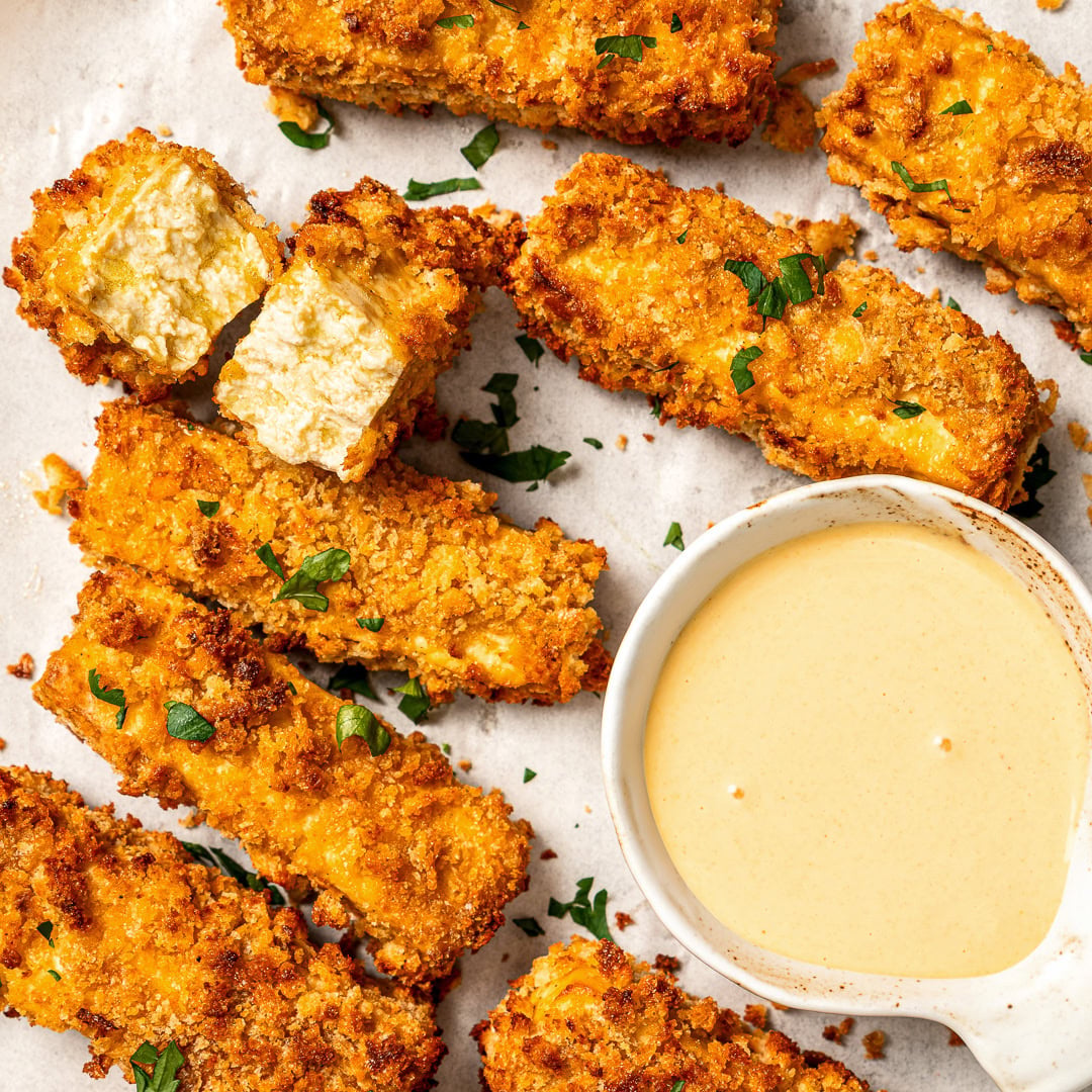 Plate of tofu chicken with sauce, with text overlay that reads vegan chicken fingers. Vegan tofu chicken fingers with bowl of sauce.