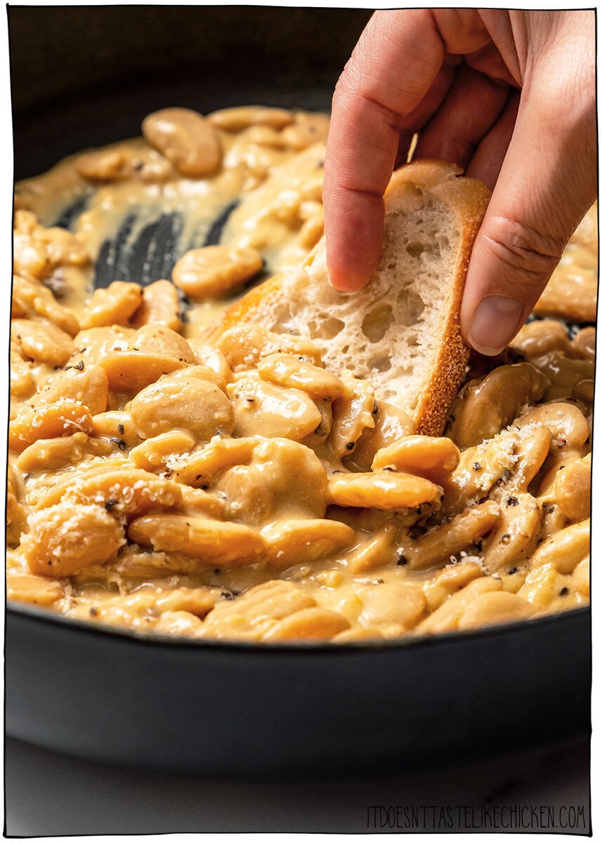 Cacio e pepe butter beans are creamy, peppery, and packed with buttery beans in a cheesy vegan sauce inspired by the classic Italian pasta! Hand dipping piece of bread into skillet of cacio e pepe butter beans.