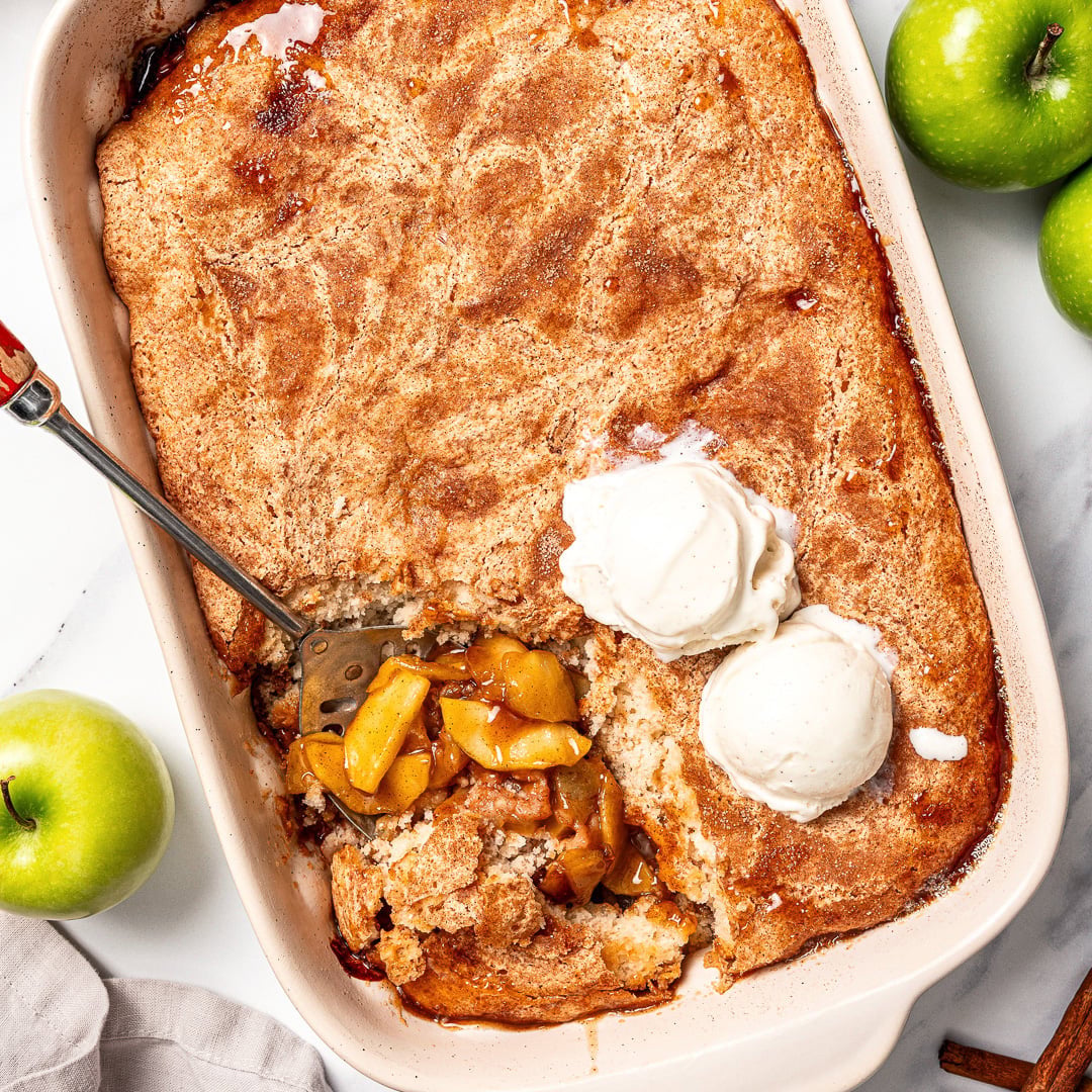 This Easy Vegan Apple Cobbler pairs tender spiced apples with a soft, buttery cake-like topping swirled with cinnamon sugar. So delicious! Spoon scooping serving of vegan apple cobbler from baking dish.